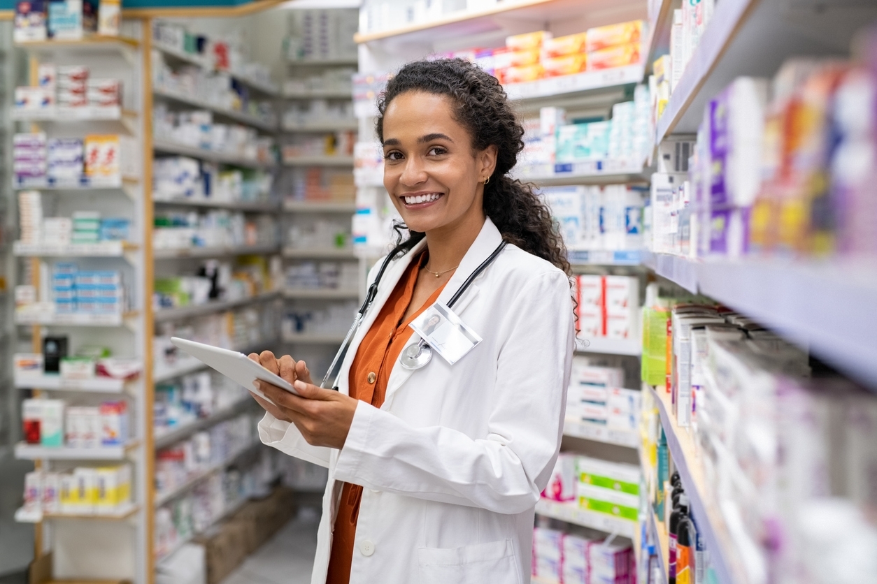 1248170454 - Happy friendly multiethnic pharmacist doing inventory in a provided and modern pharmacy while looking at camera. Portrait of smiling young doctor woman working in drugstore with digital tablet. African smiling druggist working at hospital pharmacy.
